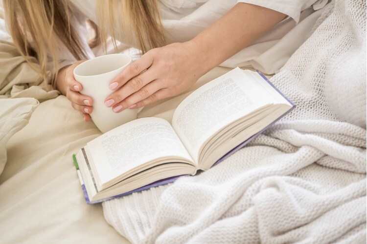 A person holding a mug and reading a book in bed, illustrating a relaxing screen-free bedtime ritual