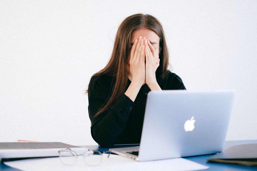 A woman covering her face in stress and fatigue while working on a laptop, illustrating digital burnout and mental exhaustion