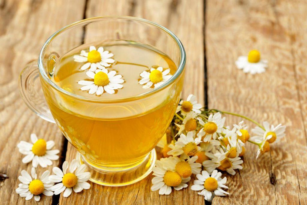 A glass cup of chamomile tea with fresh chamomile flowers floating on top and scattered around on a wooden surface, symbolizing relaxation and natural well-being A glass cup of chamomile tea with fresh chamomile flowers floating on top and scattered around on a wooden surface, symbolizing relaxation and natural well-being